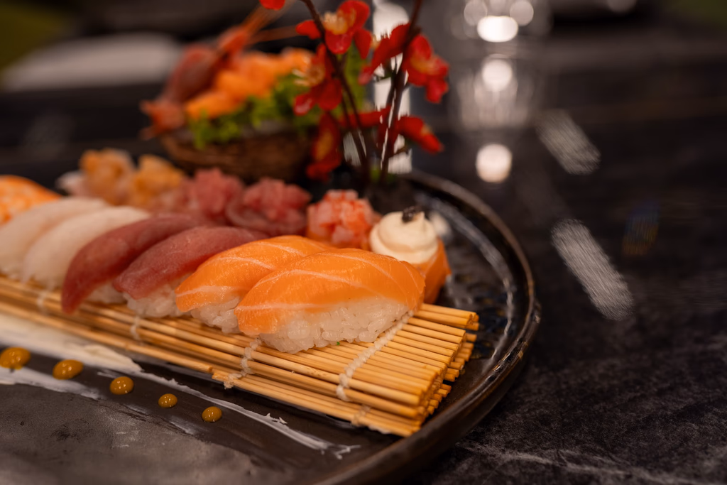 Variety of sushi pieces, including salmon and tuna nigiri, arranged on a bamboo mat with decorative red flowers on a black plate.