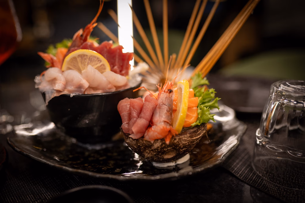 Plate of assorted sashimi with slices of raw fish, shrimp, lemon, and greens arranged with skewers on a dark table.